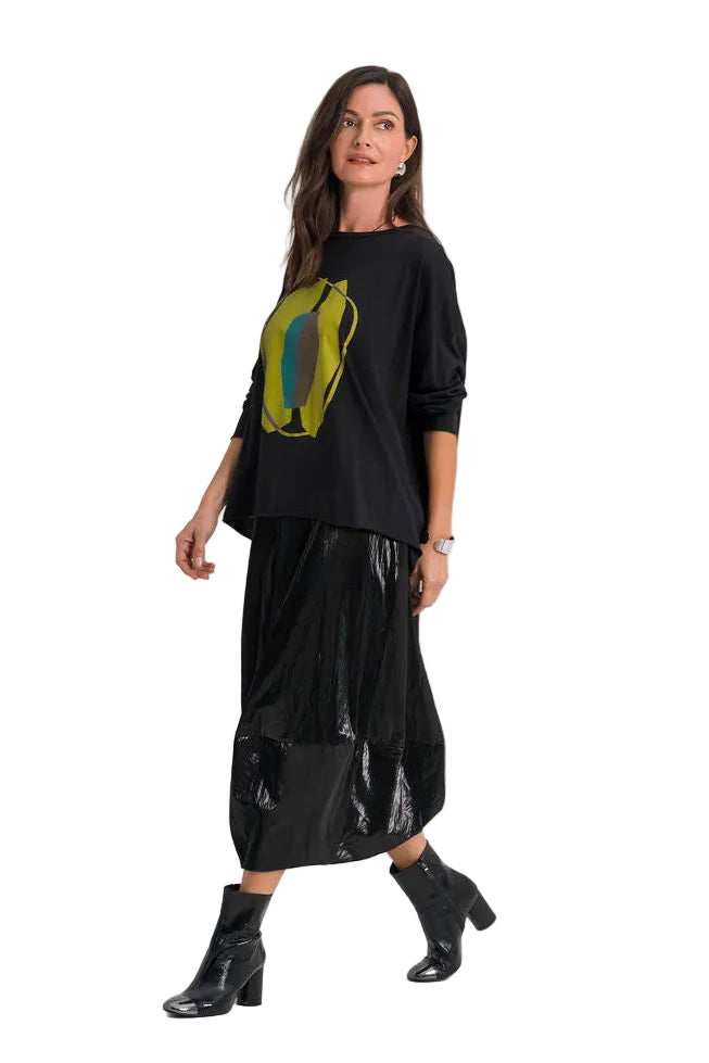 A woman modeling a 100% pima cotton black long-sleeve top with a green and yellow graphic print, paired with a black shiny midi skirt and black ankle boots, against a plain white studio background.