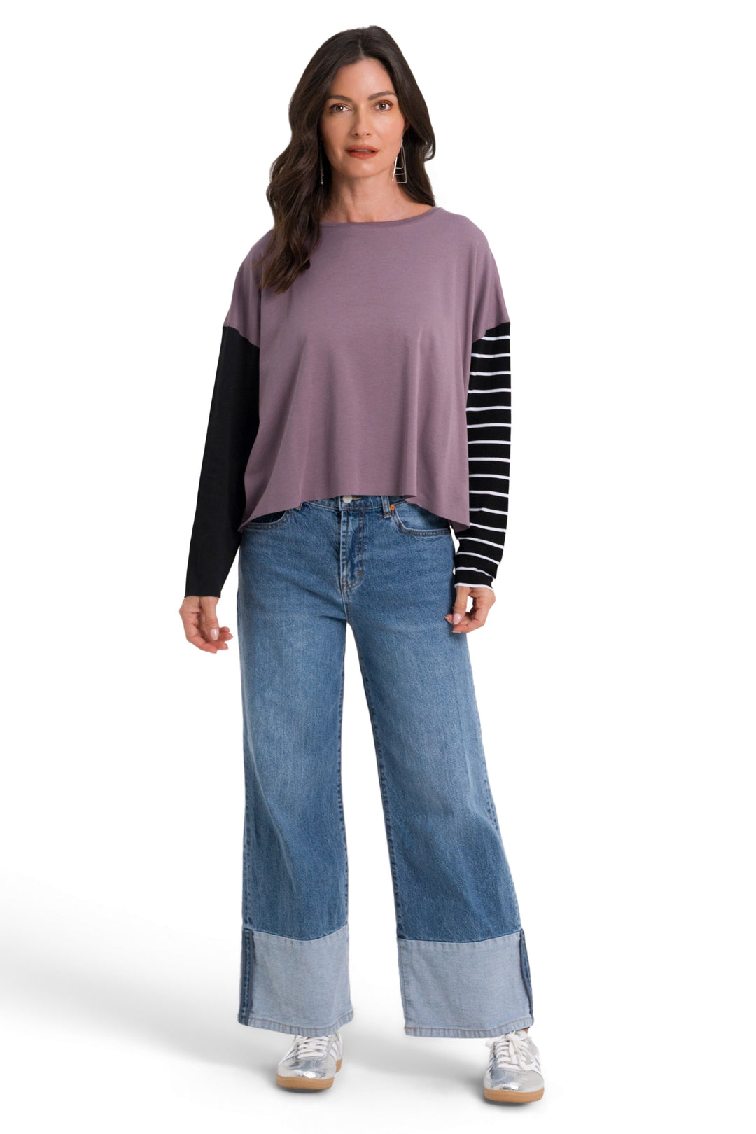 A woman modeling a 100% pima cotton colorblock long-sleeve top with a dusty purple body, black sleeves, and black-and-white striped sleeves, paired with wide-leg blue jeans and white sneakers, against a plain white background.