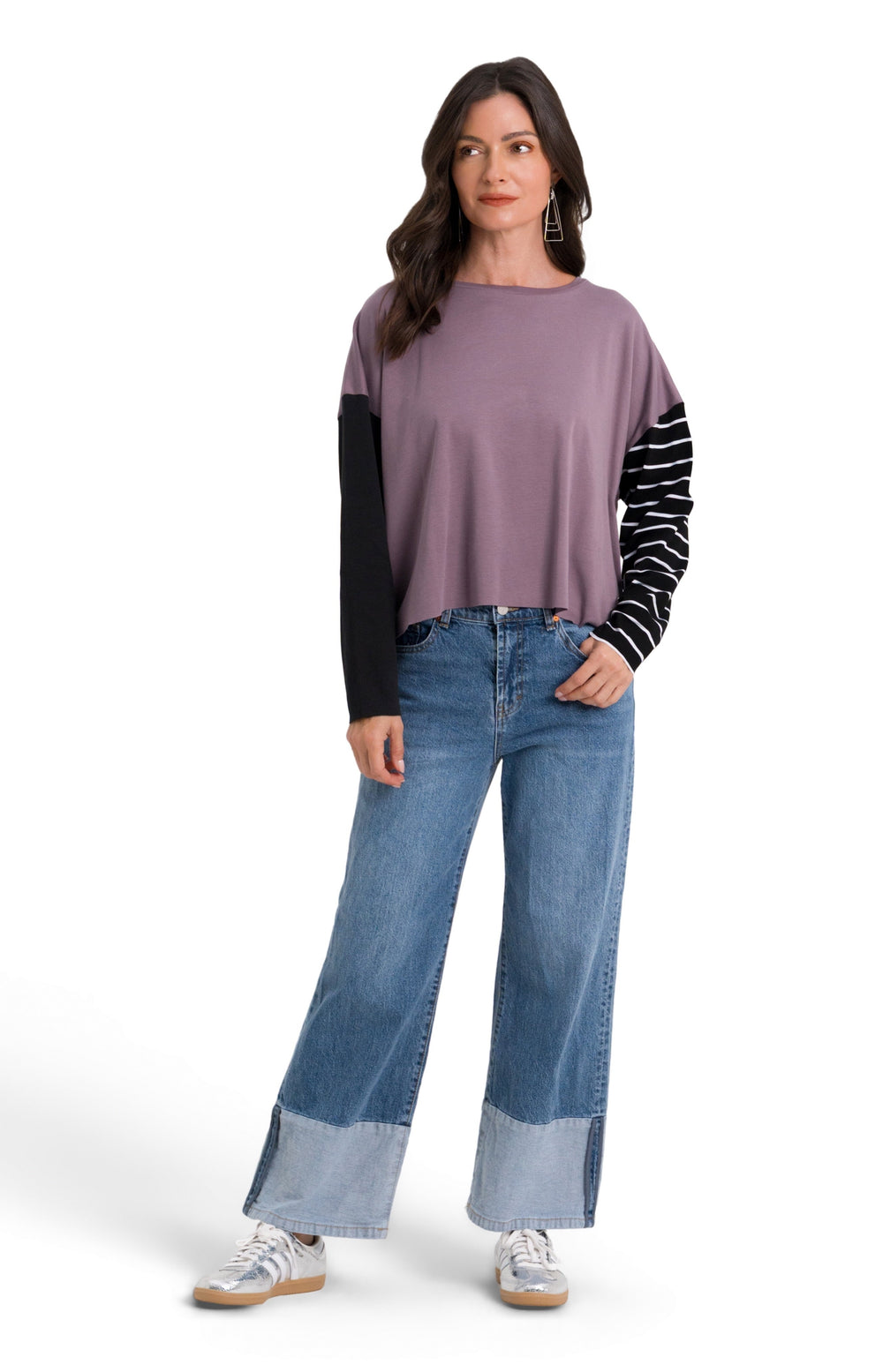 A woman modeling a 100% pima cotton color-blocked long-sleeve top with a dusty purple body, black sleeves, and black-and-white striped sleeves, paired with blue wide-leg jeans and white sneakers, on a white studio background.