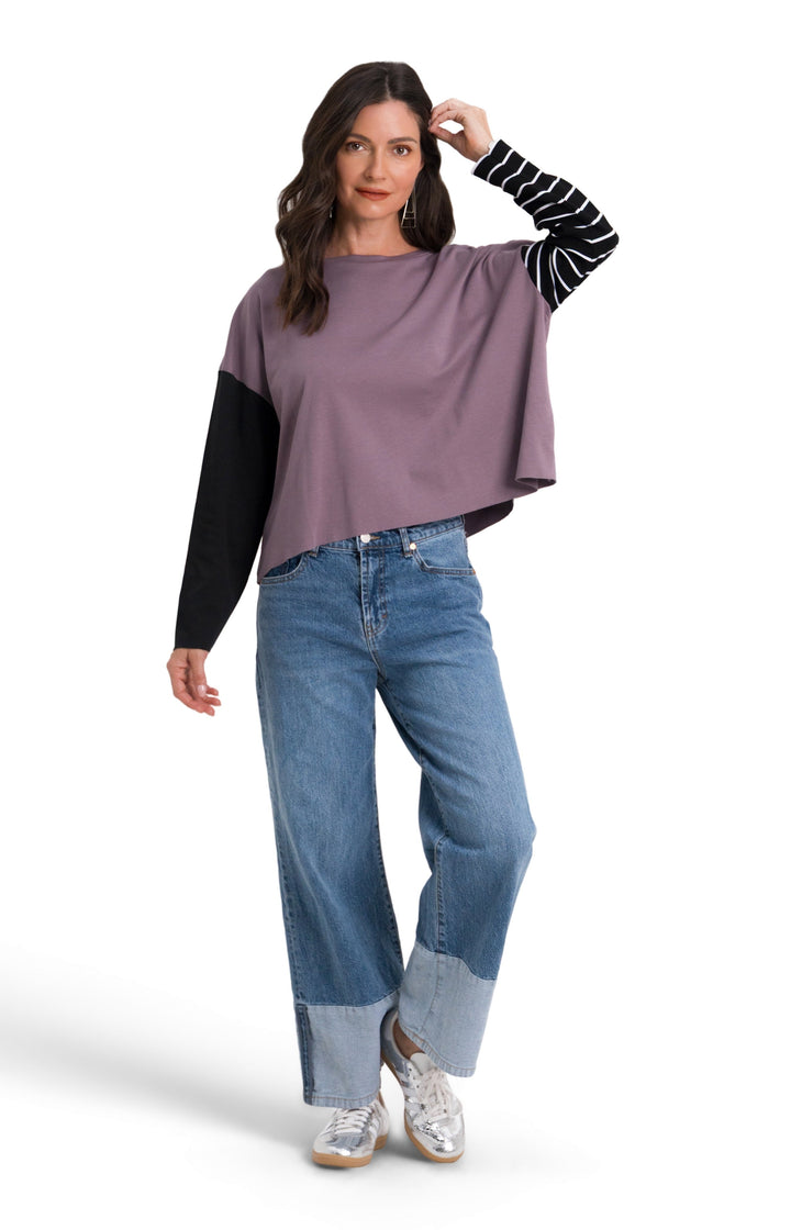 A woman modeling a 100% pima cotton color-blocked long-sleeve top with a wide purple body, black sleeves, and a black-and-white striped sleeve, paired with blue jeans and white sneakers, on a white studio background.