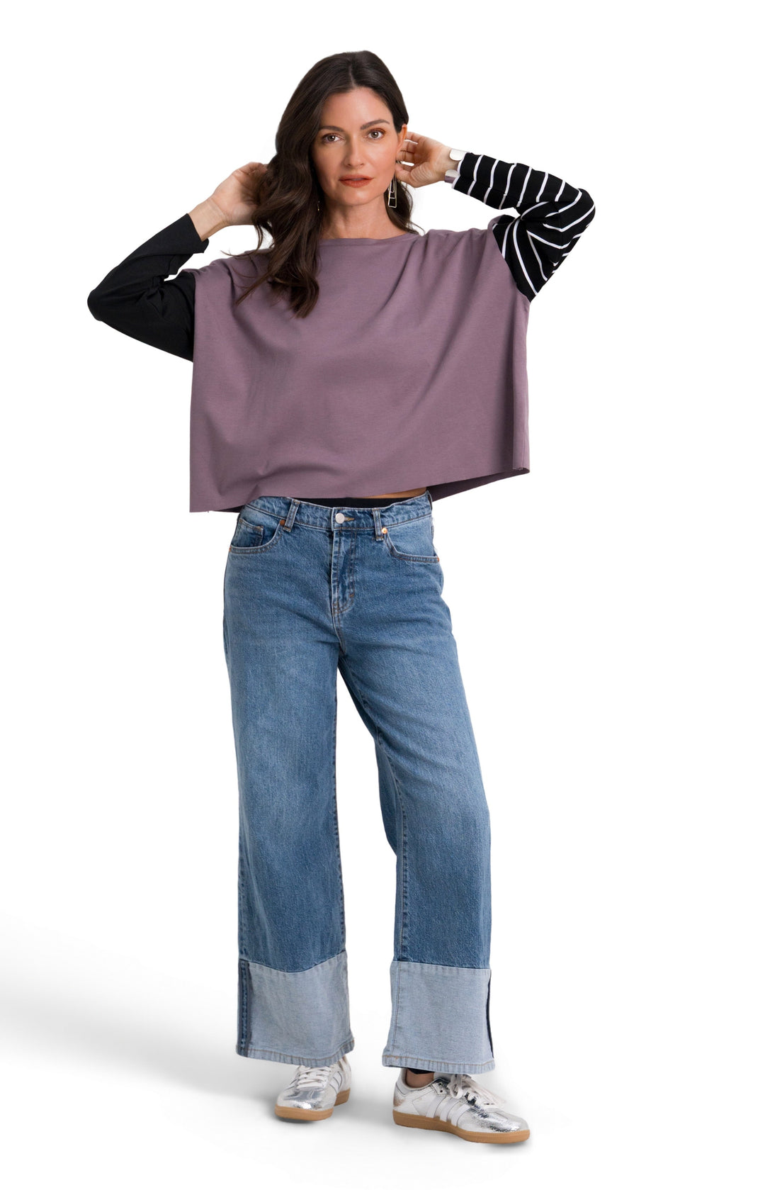 A woman modeling a loose, oversized 100% pima cotton mauve-purple top with black and white striped sleeves, paired with blue jeans and white sneakers, against a plain white background.