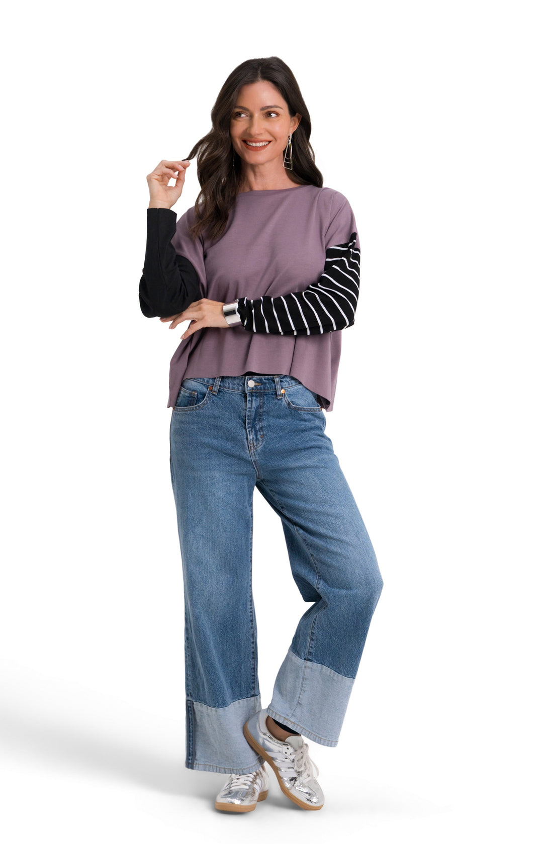 A woman modeling a 100% pima cotton mauve-purple top with black and white striped sleeves, paired with blue jeans and white sneakers, against a plain white background.