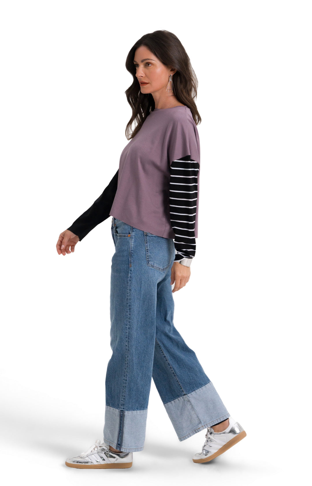 A woman walking in profile wearing a 100% pima cotton mauve-purple top with black and white striped sleeves, blue jeans with rolled cuffs, and white sneakers, set against a plain white background.