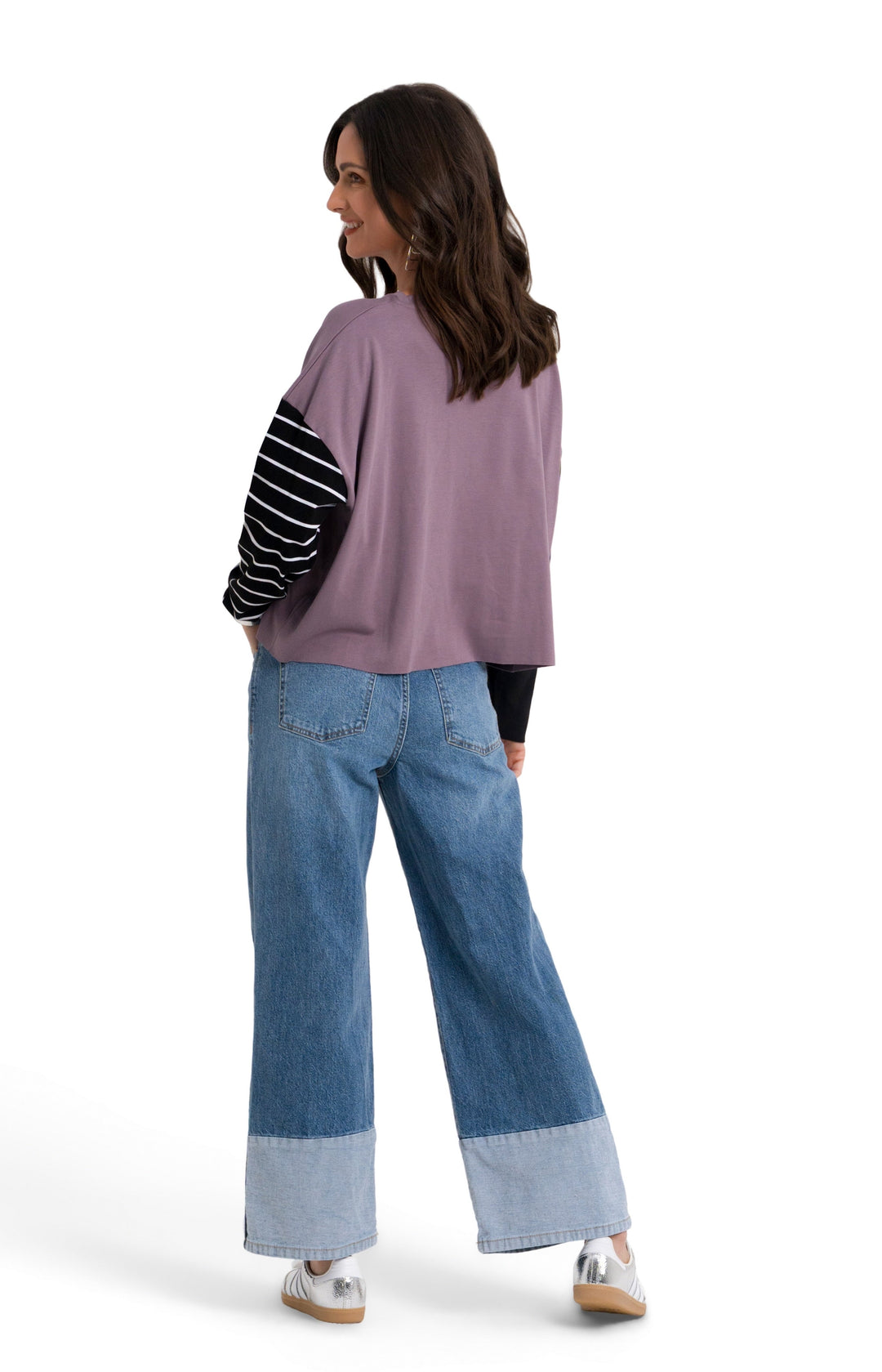 A woman is shown from behind wearing a 100% pima cotton lavender-purple cropped top with black and white striped sleeves, blue wide-leg jeans with cuffed hems, and white sneakers, set against a plain white studio background.
