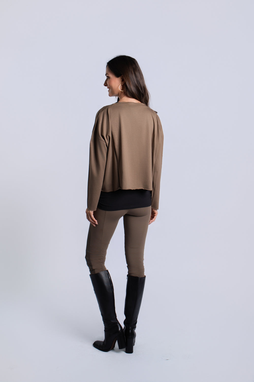 A woman is shown from the back wearing a turtle-brown draped long-sleeve pima cotton top, matching taupe-brown pants, and black knee-high boots against a light gray studio background.