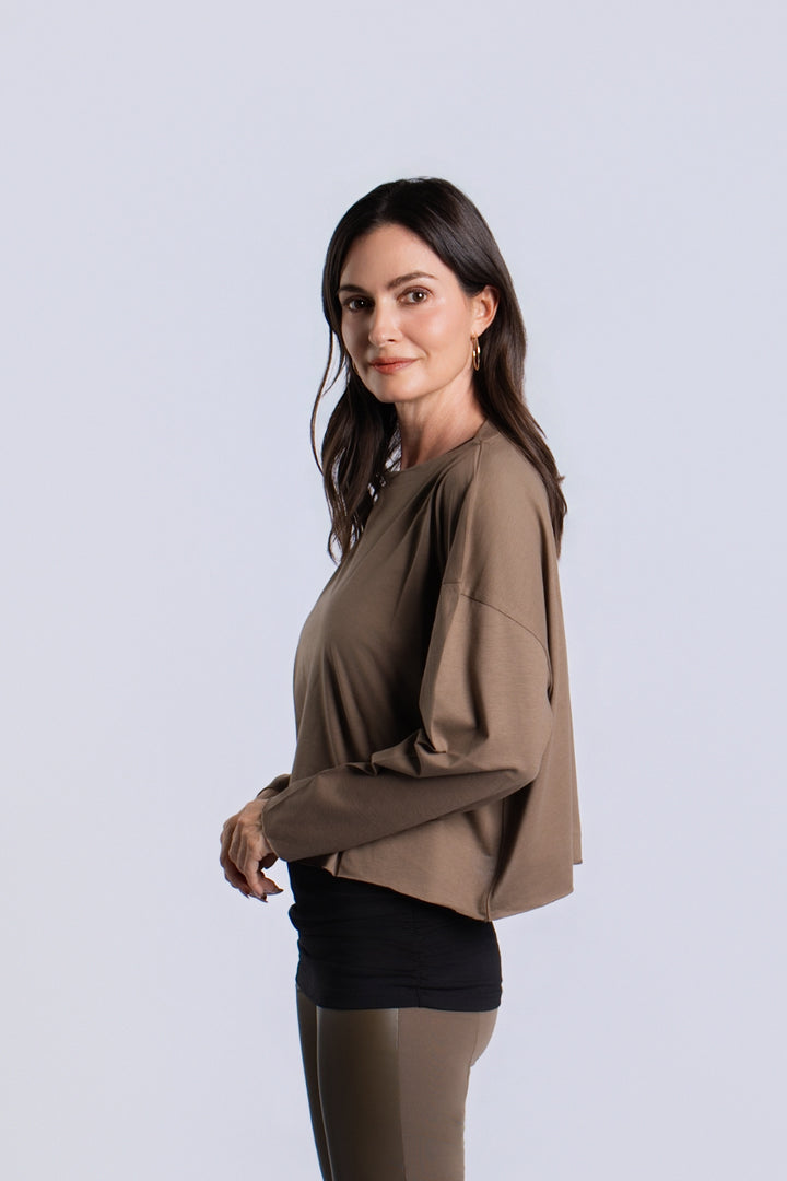 A woman modeling a cropped, long-sleeve turtle-brown pima cotton top with a crew neckline, paired with black shorts and olive leggings, against a light gray studio background.