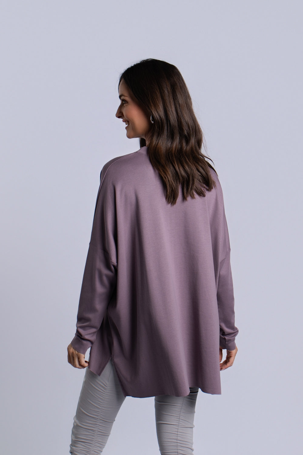 A woman with long brown hair is shown from the back wearing a loose, long-sleeve tunic top in a dusty mauve-purple color, paired with light gray leggings, against a solid light gray studio background.