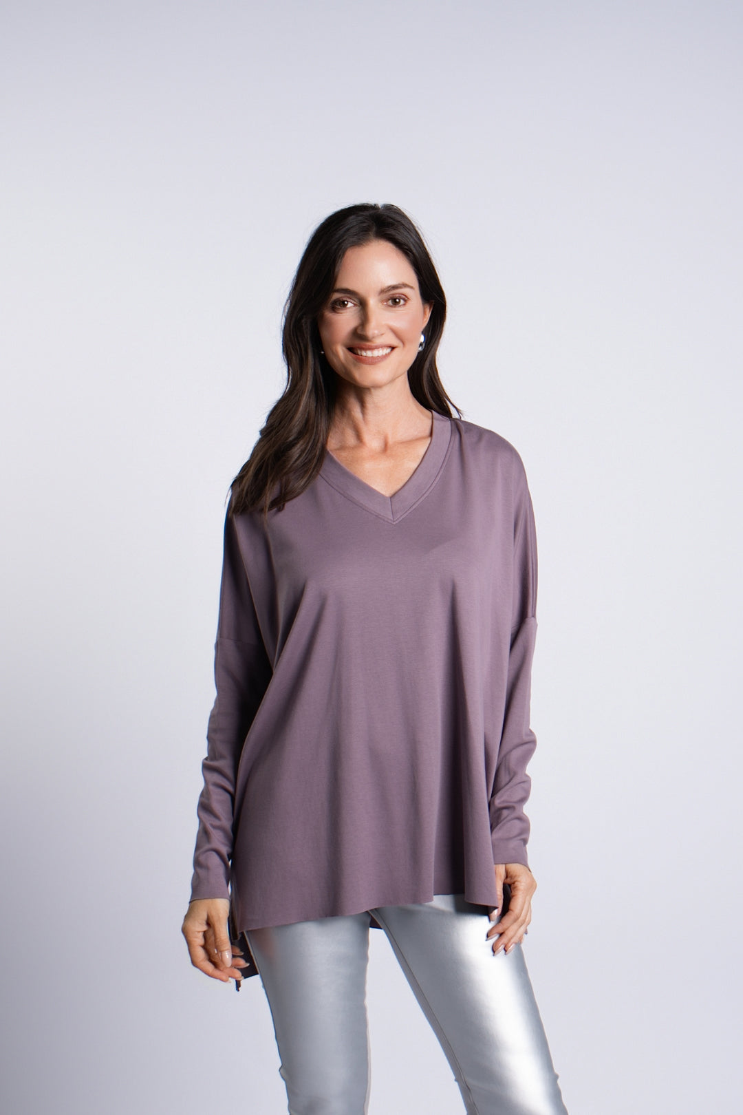 A woman modeling a long-sleeve pima cotton V-neck top in a muted dusty purple color, paired with metallic silver pants, against a light gray studio background.