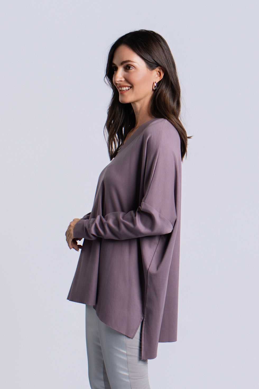 A woman wearing a loose, long-sleeve pima cotton V-neck top in a muted mauve-purple color, paired with light gray pants, photographed against a solid light gray studio background.