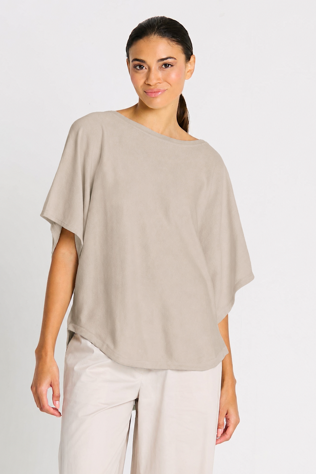 Woman wearing a beige putty Planet one-shoulder top and white pants on a light gray background.