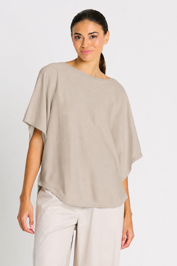 Woman wearing a beige putty Planet one-shoulder top and white pants on a light gray background.