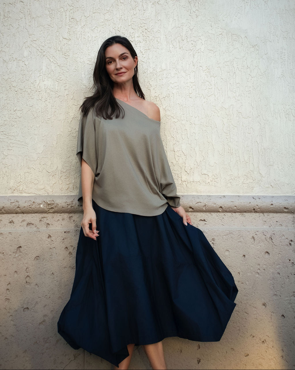 A woman posing in a loose, off-the-shoulder taupe top and a dark navy blue skirt against a textured beige stucco wall.