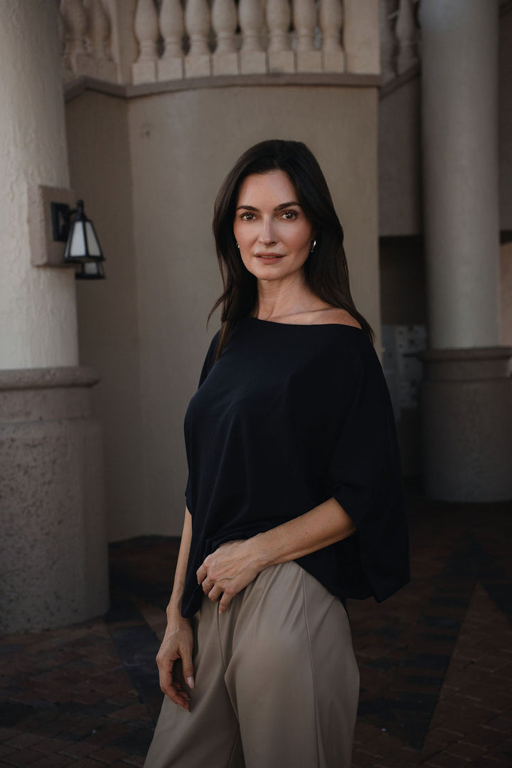 A woman with dark hair stands in a stone corridor wearing a black off-the-shoulder top and beige wide-leg pants.