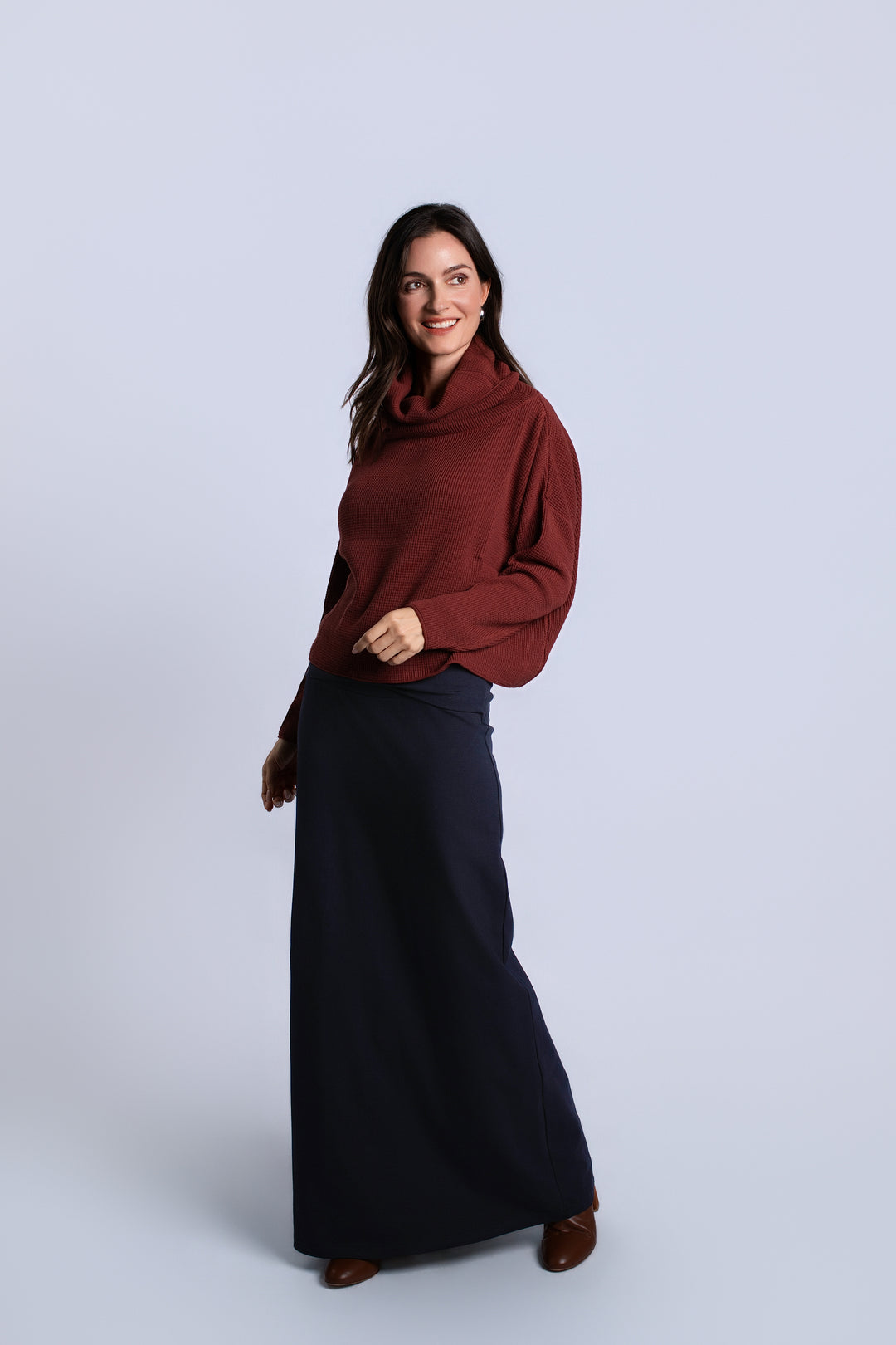 A woman modeling a dark red cowl-neck sweater and a long navy skirt against a light gray studio background.