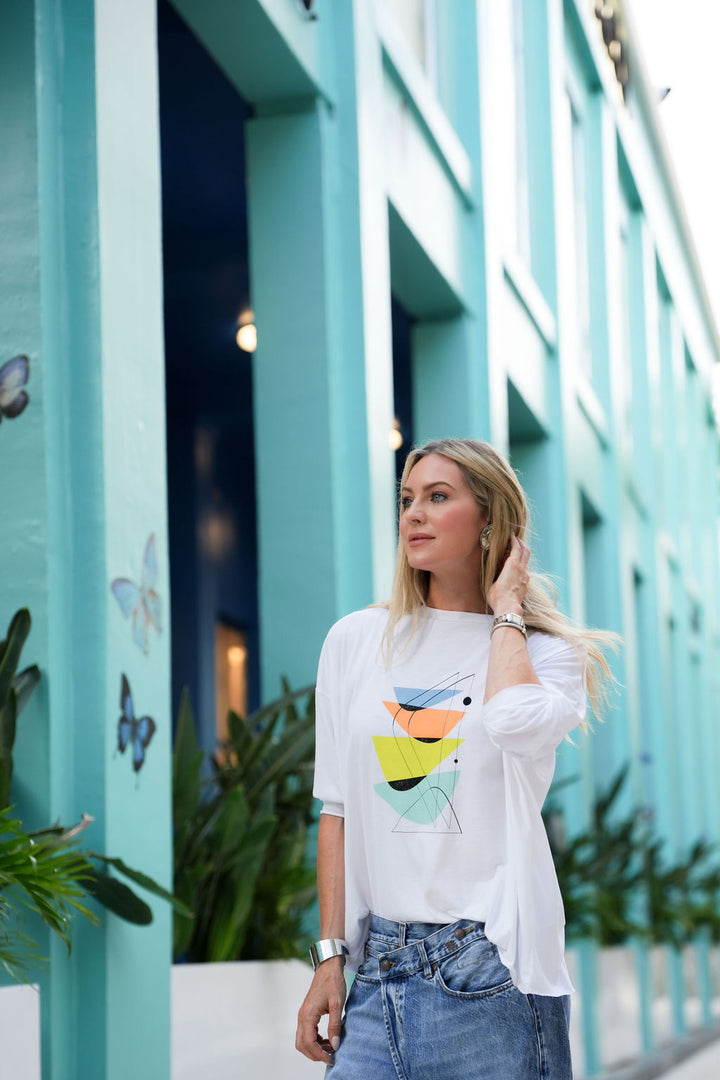 Woman wearing a white pima cotton shirt with a colorful design in front of a turquoise building.