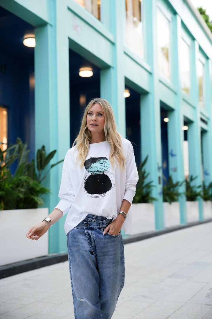 Woman wearing a white pima cotton shirt with a graphic design and blue jeans standing in front of a teal building.