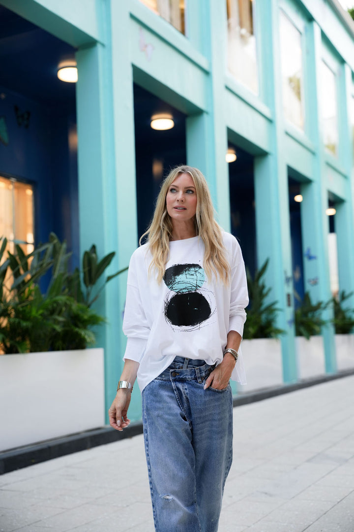 Woman wearing a white pima cotton shirt with a graphic design and blue jeans standing in front of a turquoise building.