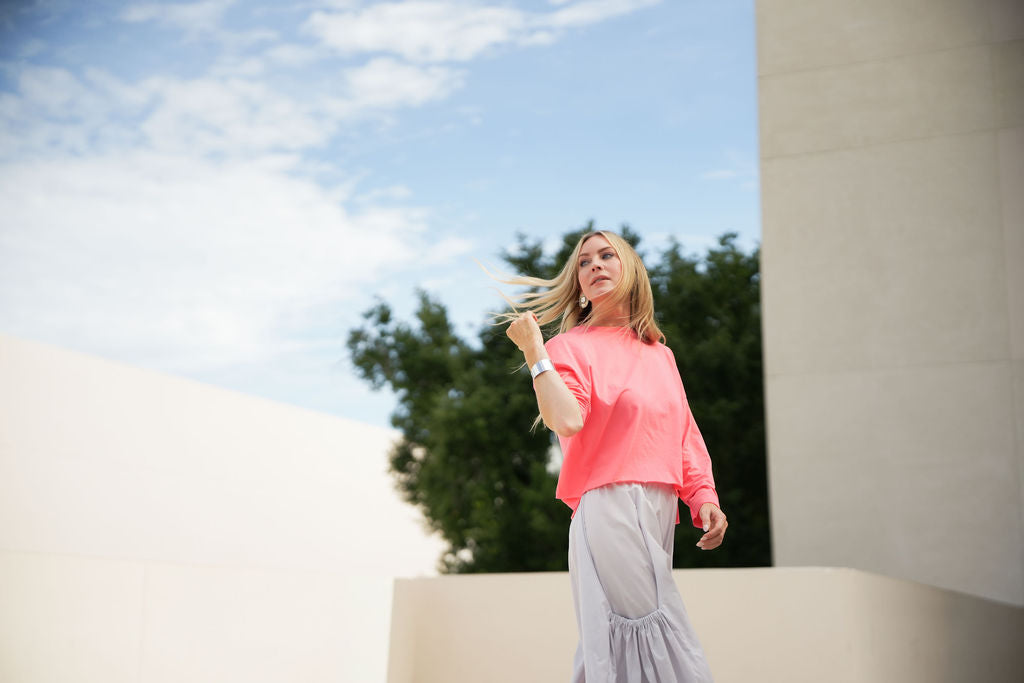 Woman in a coral pink pima cotton boxy t-shirt and gray pants standing outdoors with trees and blue sky in the background.