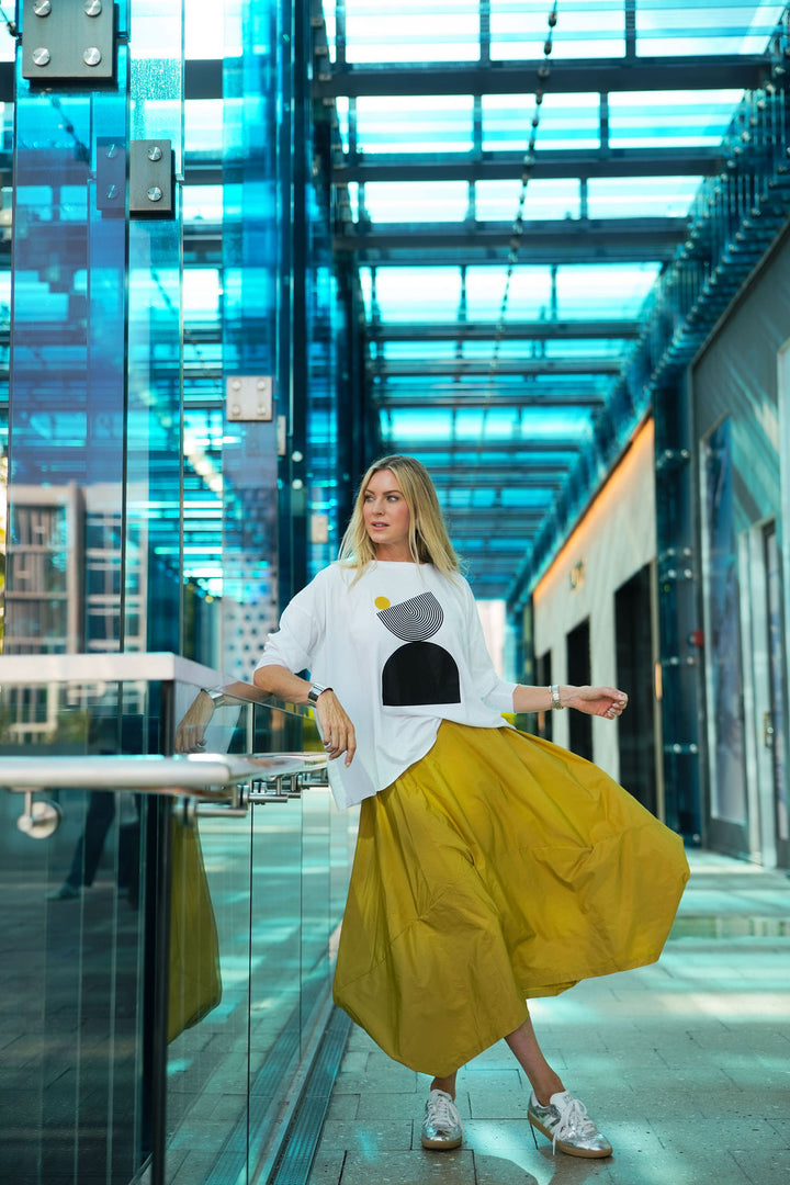 Woman in a white pima cotton boxy t-shirt with a graphic design and yellow skirt standing in a modern building.