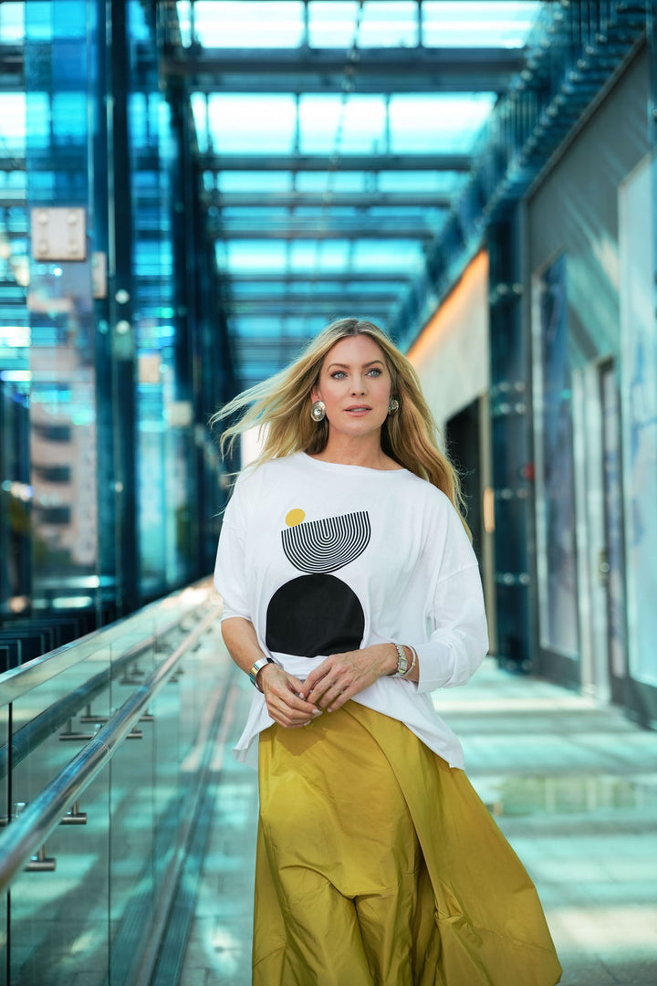 Woman in a pima cotton white boxy t-shirt with a black abstract design and yellow pants standing in a modern building.