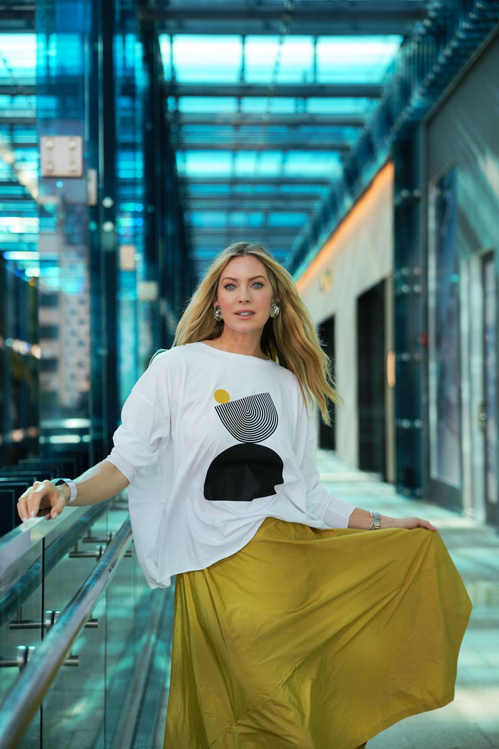 Woman in a white pima cotton boxy t-shirt with a black and yellow design and a yellow skirt standing in a modern indoor setting.
