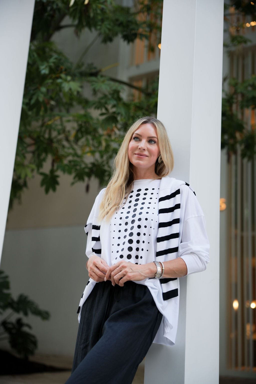 Woman wearing a white pima cotton shirt and cardigan with black polka dots and stripes, standing against a white column with greenery in the background.