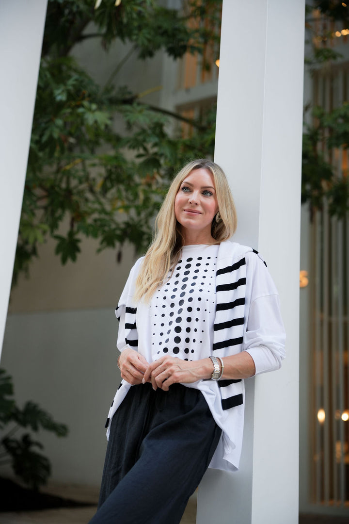 Woman wearing a white pima cotton shirt and cardigan with black polka dots and stripes, standing against a white column with greenery in the background.