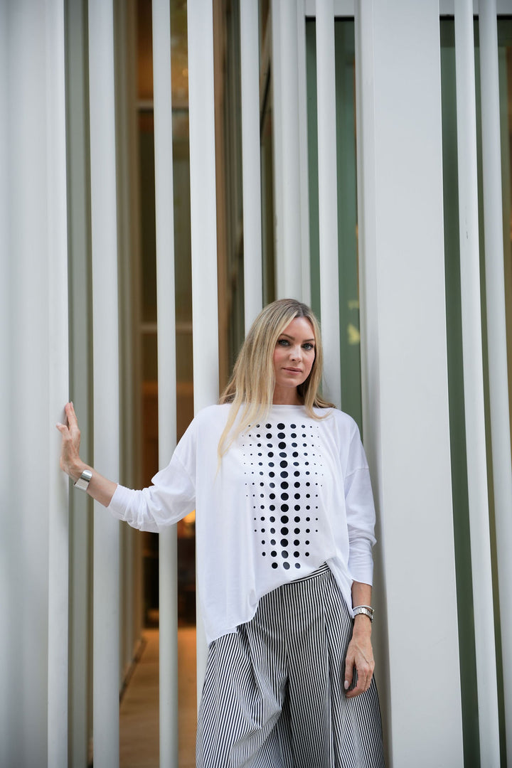 Woman wearing a white pima cotton top with black patterns and gray pants standing against a modern architectural background.