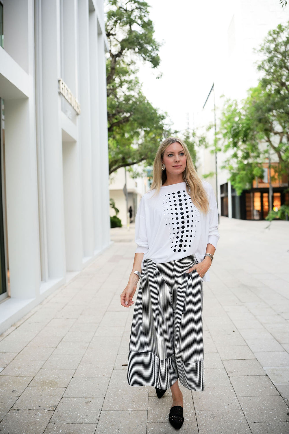Woman wearing a white blouse with black pattern and gray wide-leg pants walking outdoors.