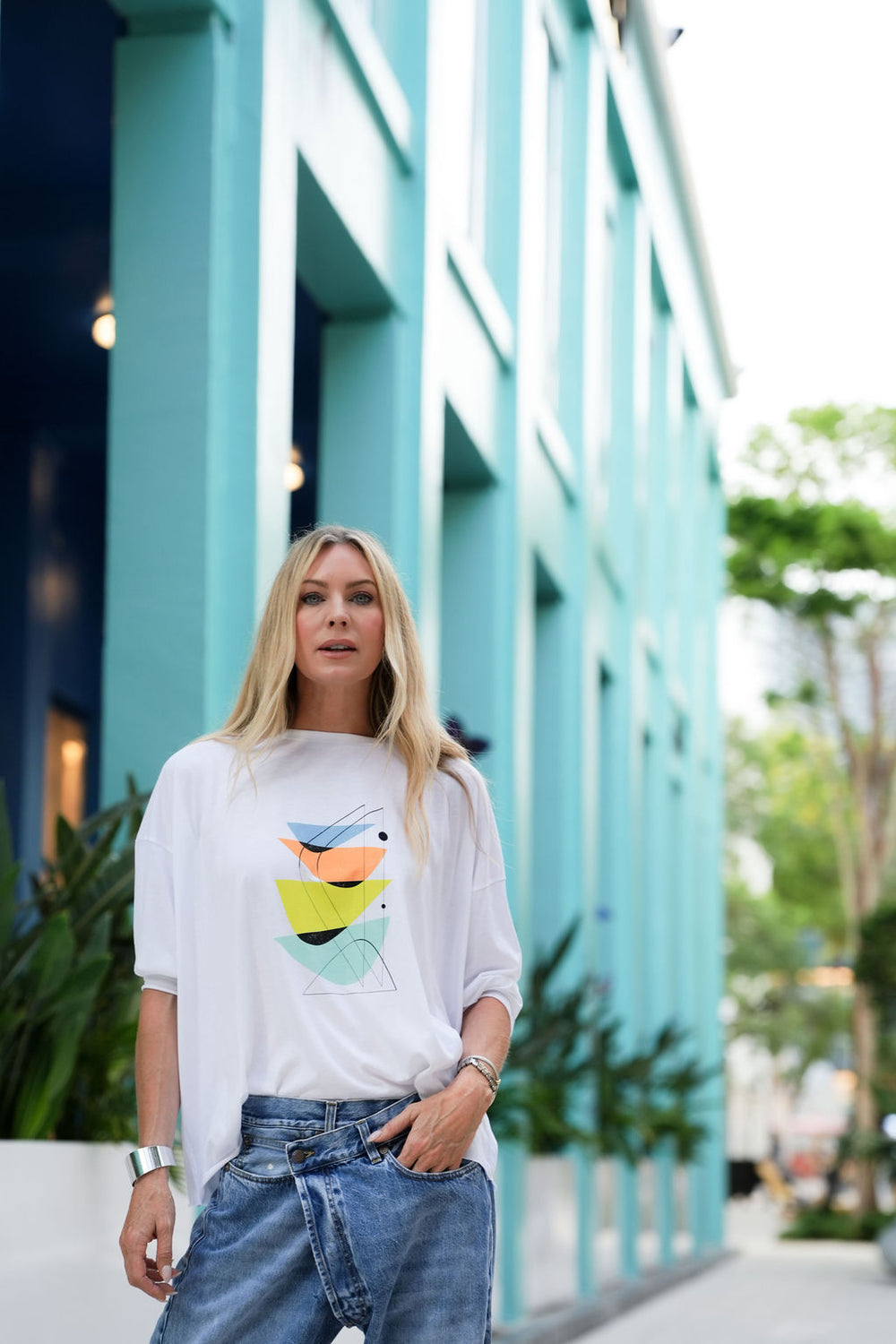 Woman wearing a white pima cotton t-shirt with a colorful graphic design in front of a teal building.