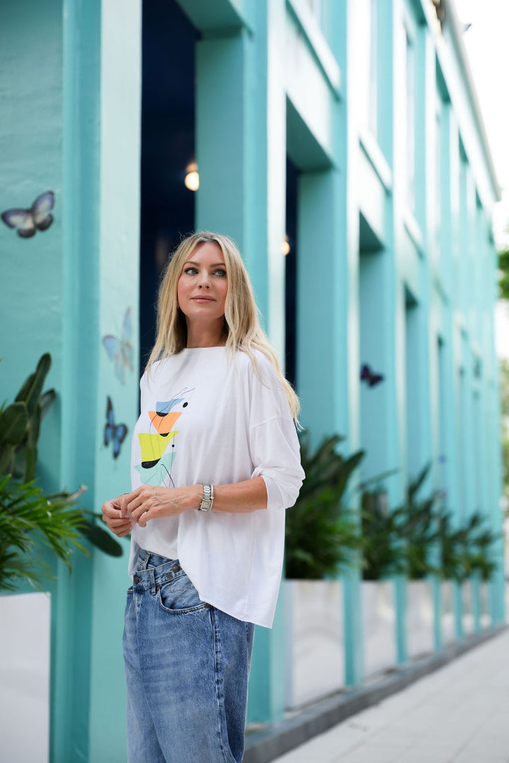 Woman in a white pima cotton shirt with a colorful design and blue jeans standing in front of a teal building.