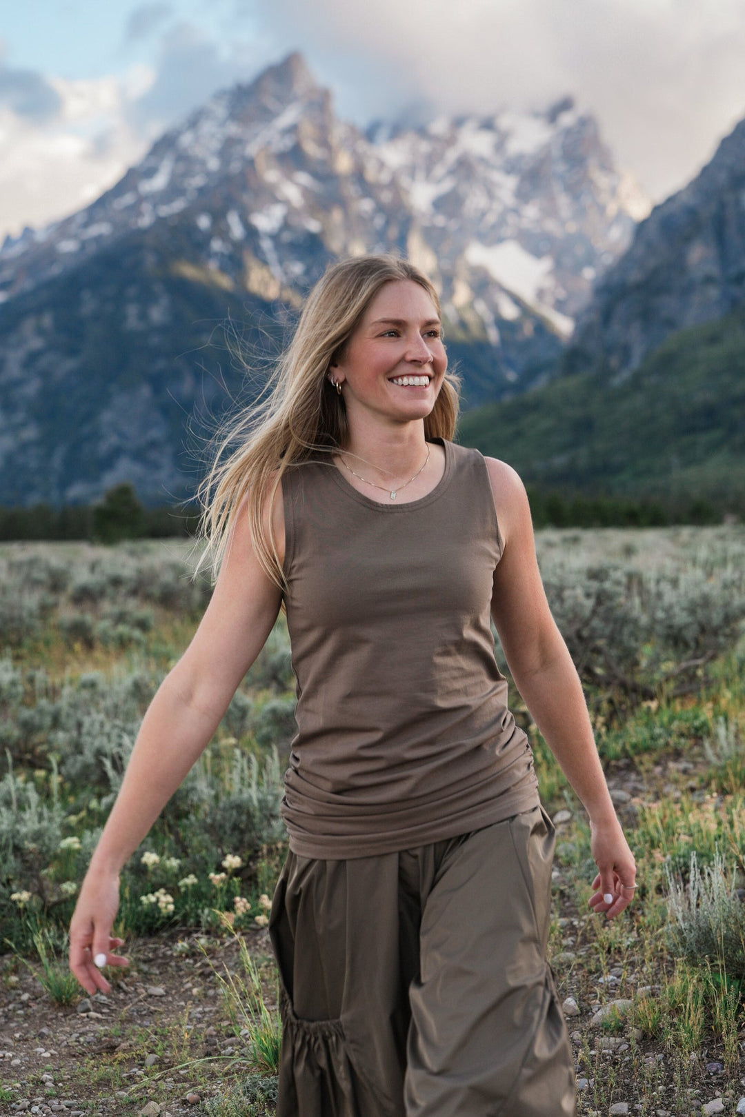A woman smiling and walking outdoors in a mountain meadow with snow-capped peaks in the background, wearing a cotton lycra brown sleeveless top and matching brown cargo pants.