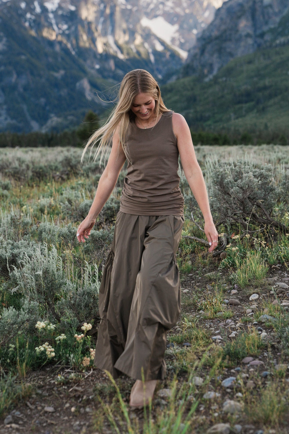 A woman walking barefoot on a dirt path in a grassy mountain meadow, wearing a cotton lycra brown sleeveless top and matching wide-leg pants, with snow-capped mountains in the background.