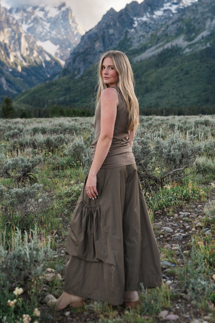 A woman stands in a grassy mountain meadow with snow-capped peaks and a blue sky behind her, wearing a sleeveless taupe-brown cotton lycra tank top and tan boots.