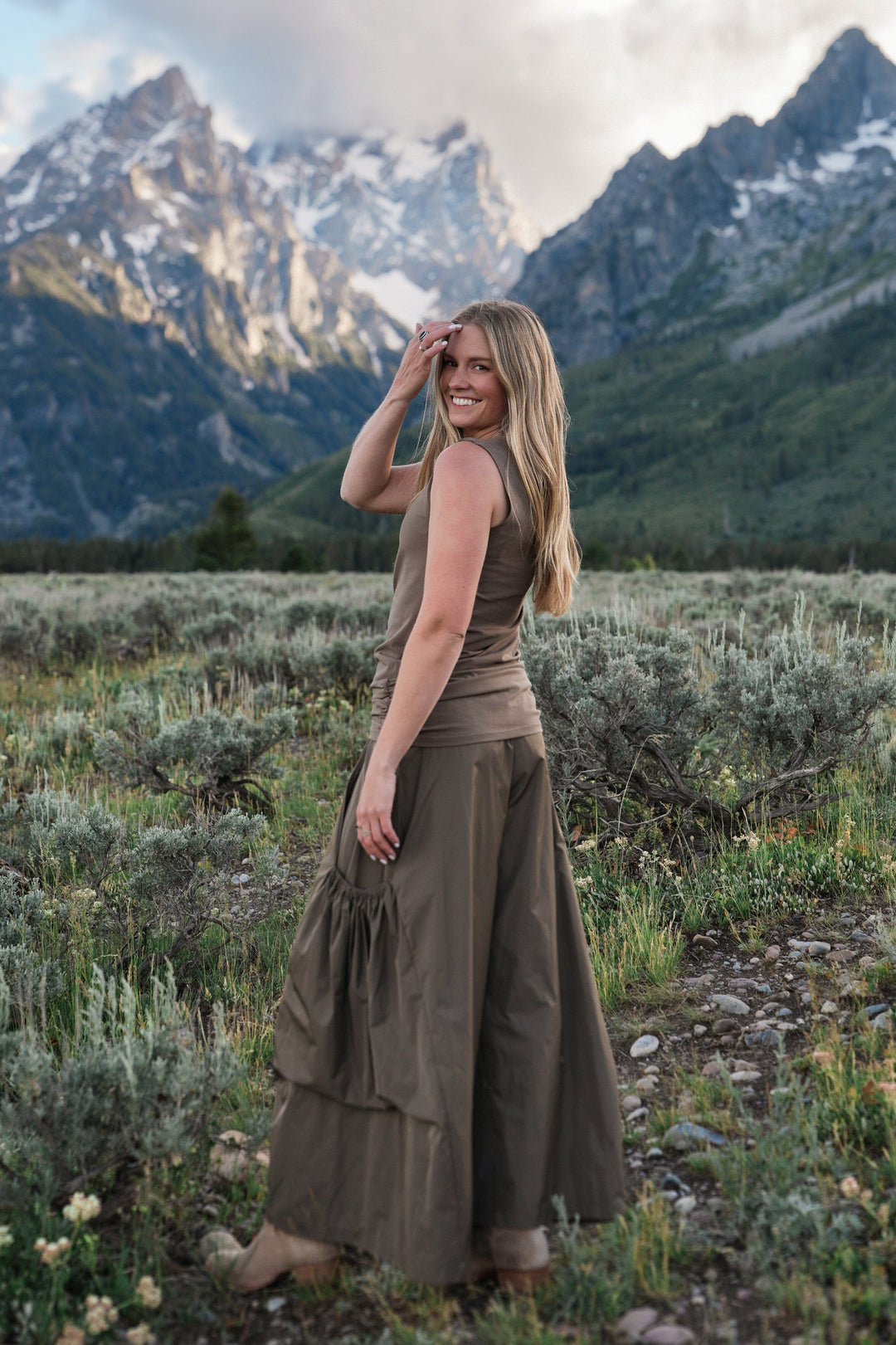 A woman stands in a grassy mountain meadow with rugged peaks and a cloudy sky behind her, wearing a sleeveless taupe-brown cotton lycra tank top. The scene is outdoors in a natural landscape, so the subject type is classified as Lifestyle.