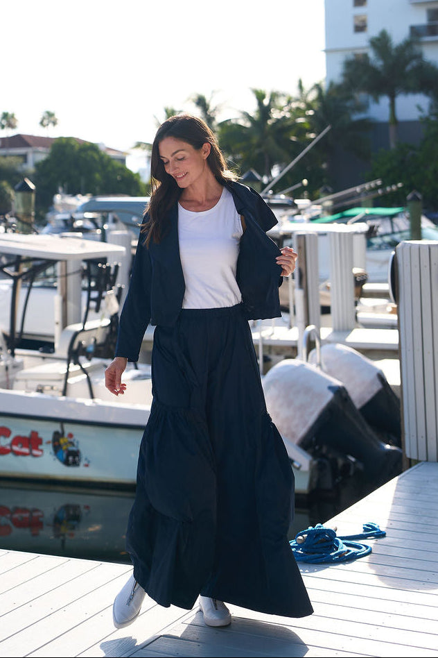 Woman in a midnight big pocket pants and a white pima cotton shirt standing on a dock with boats and buildings in the background.