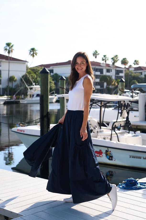 Woman standing on a dock by a marina with boats and buildings in the background wearing white shoes and midnight navy big pocket pants from PLANET by Lauren G.