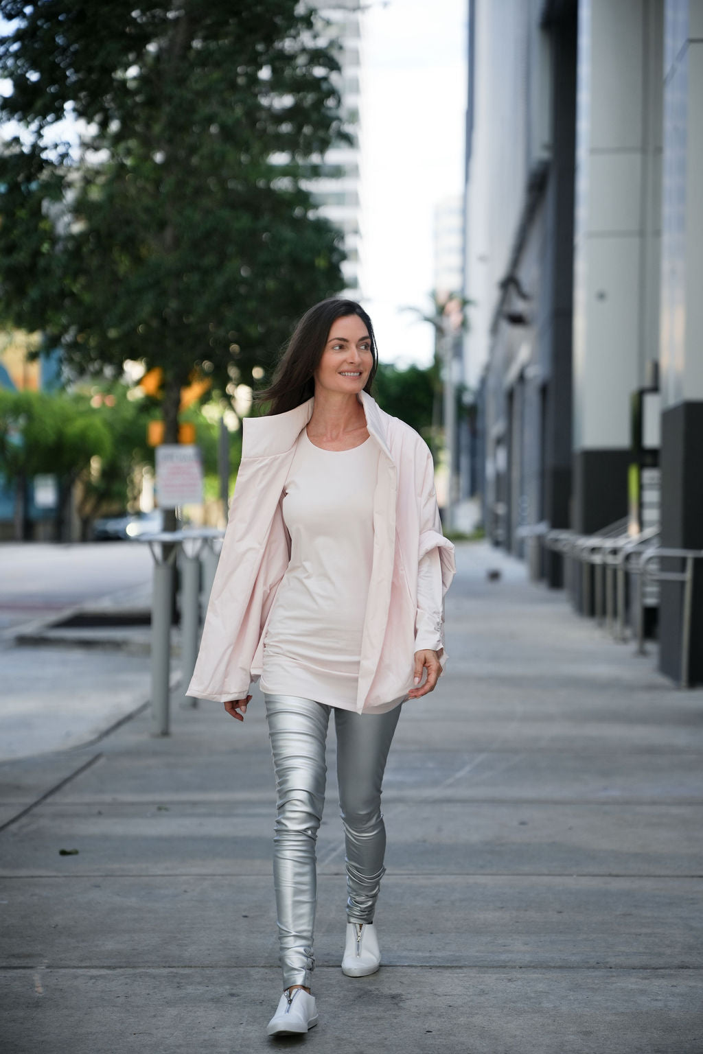 Woman walking on a city street wearing a light pink jacket, white top, and silver leggings.