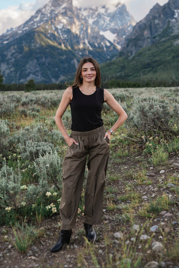 A woman stands in a grassy mountain landscape wearing a black sleeveless tank top and loose brown pants, with snow-capped peaks and clouds in the background.
