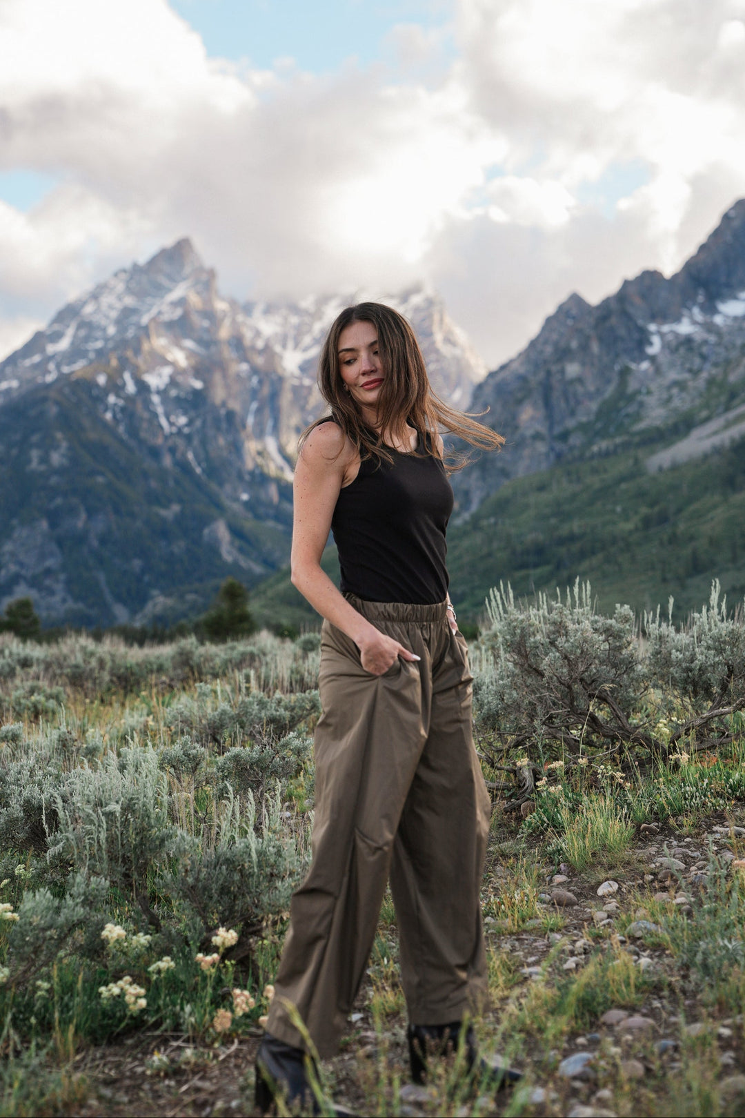 A woman stands outdoors in a mountain landscape wearing a black sleeveless top and loose olive-brown cargo pants, with snow-capped peaks and blue sky behind her.