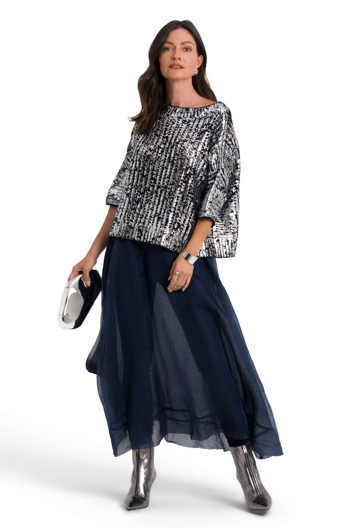 A woman modeling a three-quarter sleeve top with a black and white textured pattern, paired with a long navy blue skirt and metallic silver boots, holding a small black clutch against a plain white background.