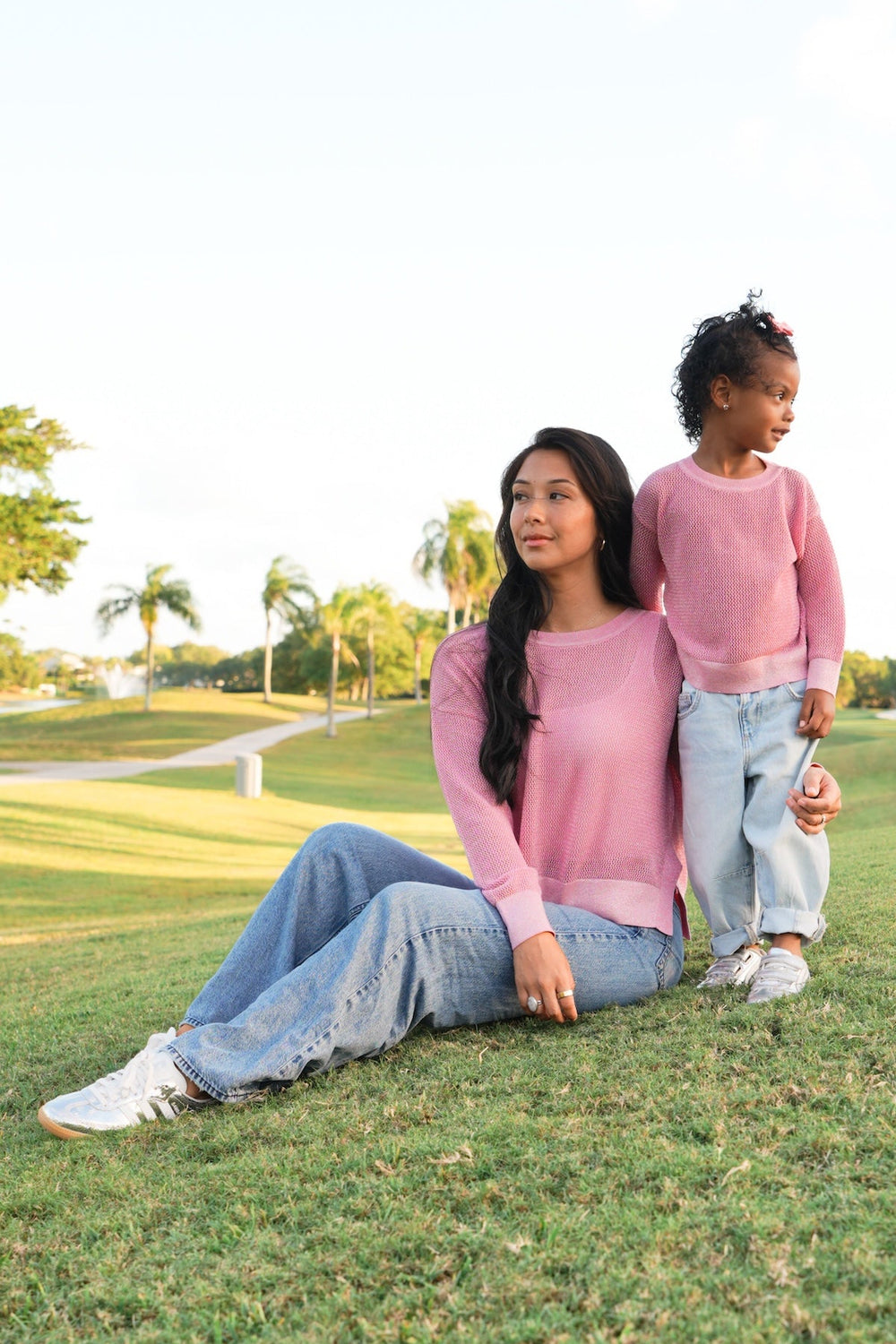 Woman and child sitting on a grassy field with trees in the background in pink pima cotton sweaters
