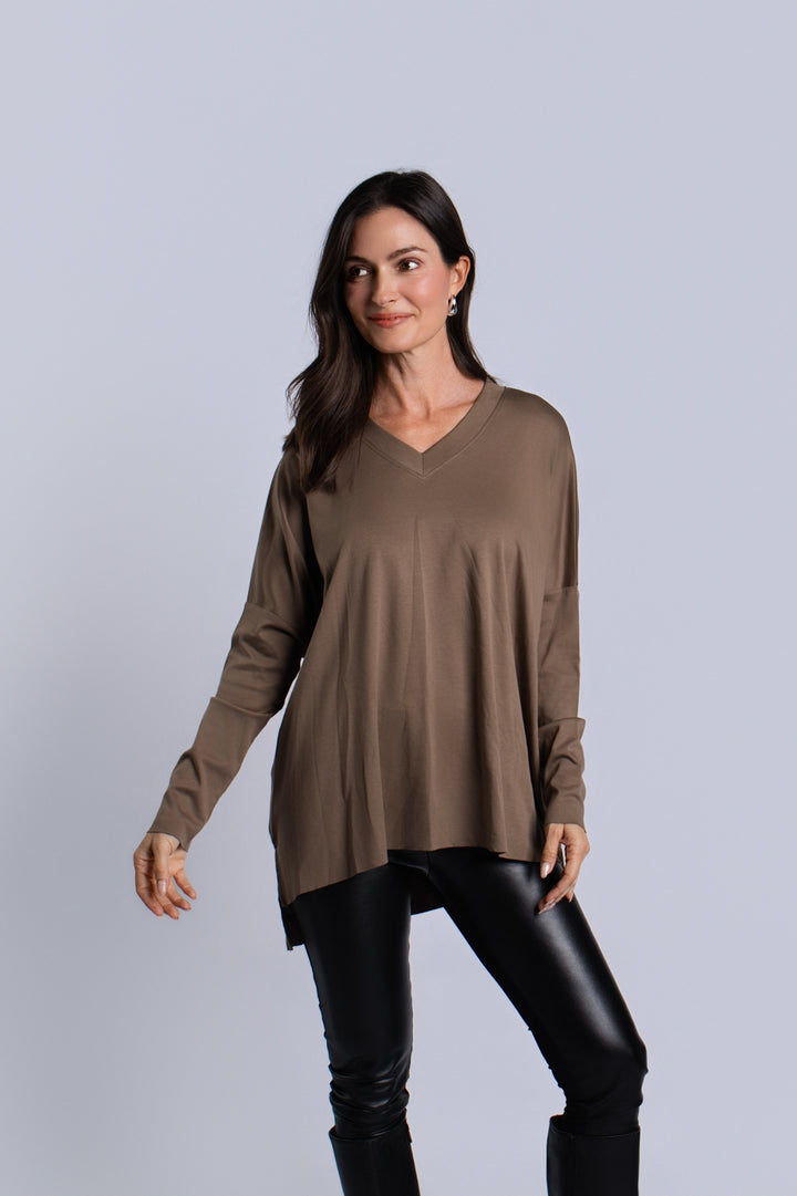 A woman modeling a turtle brown long-sleeve pima cotton V-neck tunic top with black faux-leather pants, standing against a solid light gray studio background.