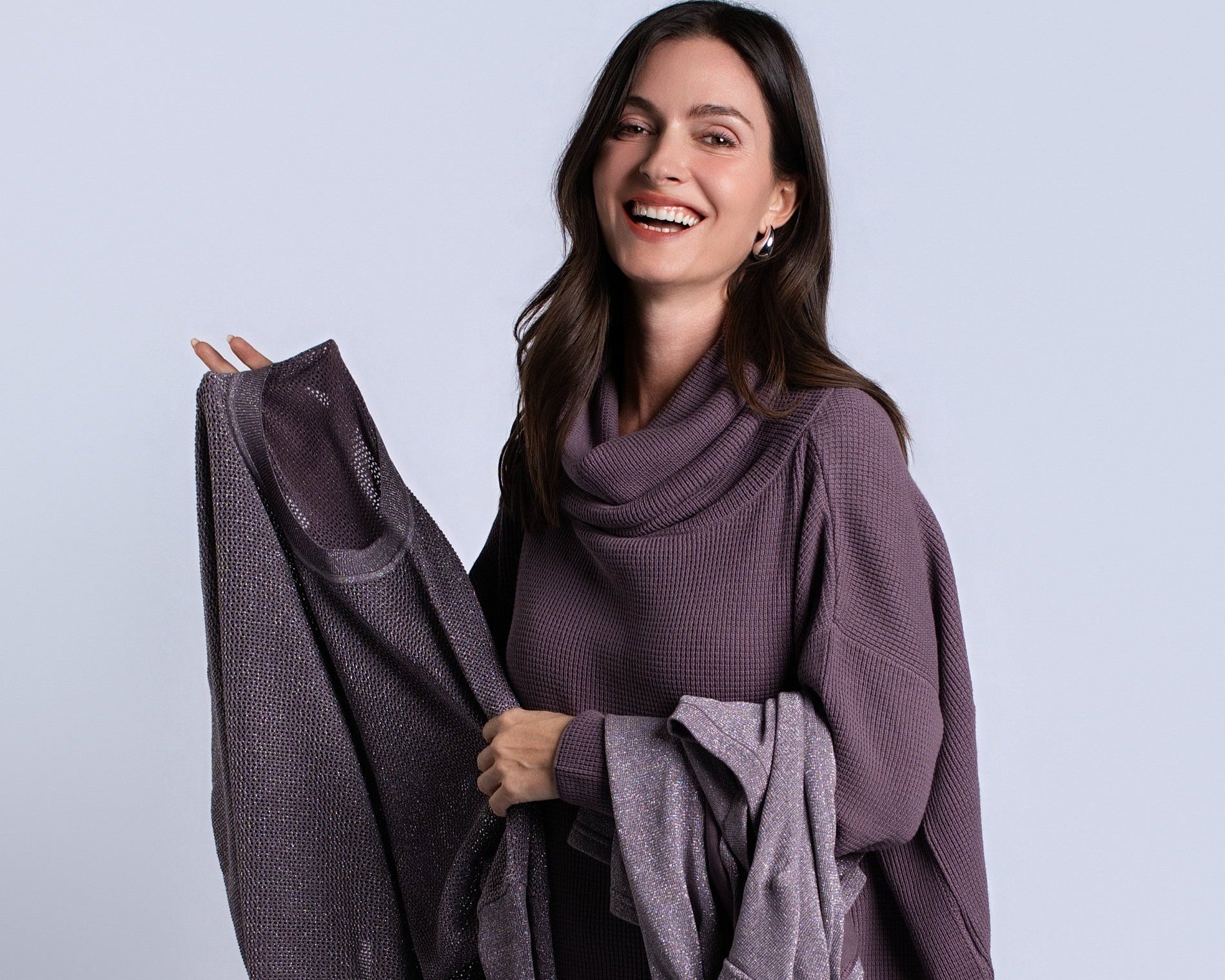 A woman smiling and holding up a matching knit sweater and metalic sweater set in an amethyst purple color, posed against a solid light gray studio background.