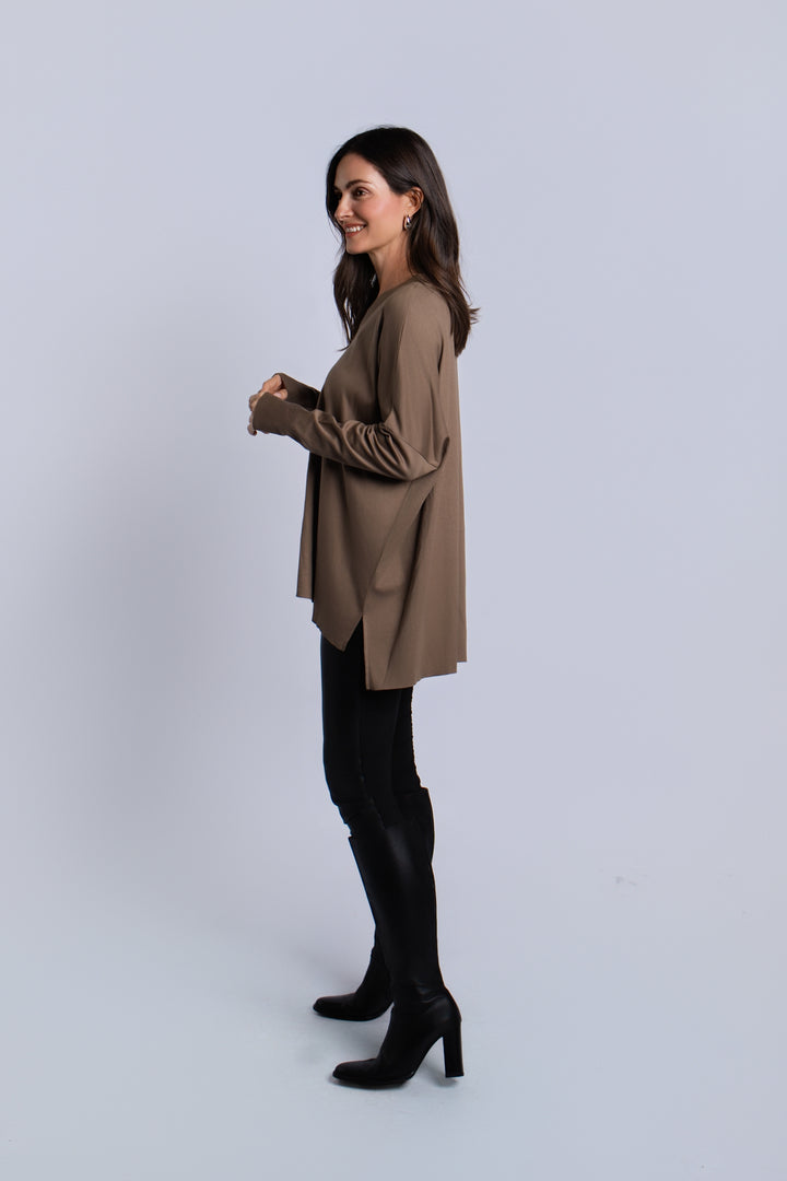 A woman modeling a loose-fitting turtle brown long-sleeve pima cotton V neck top, paired with black pants and black heeled boots, standing against a light gray studio background.