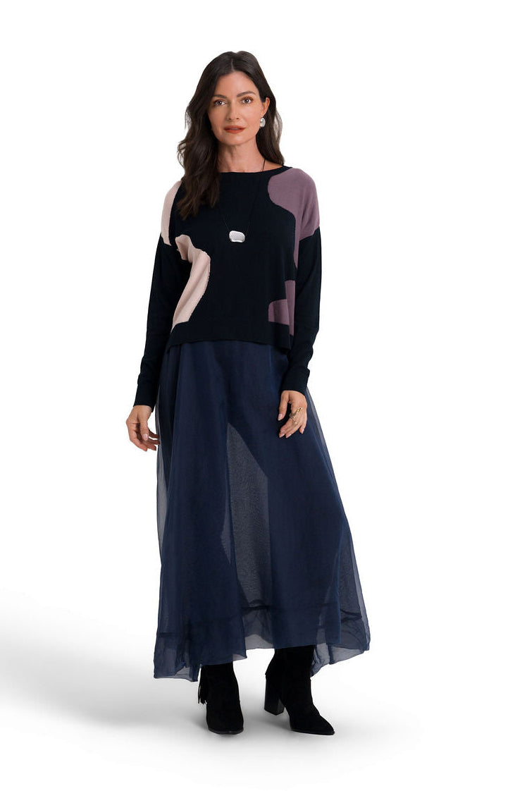 A woman modeling a long-sleeve pima cotton sweater and matching midi skirt set. The color-blocked top features navy, black, dusty purple, and beige sections, paired with a sheer navy midi skirt.