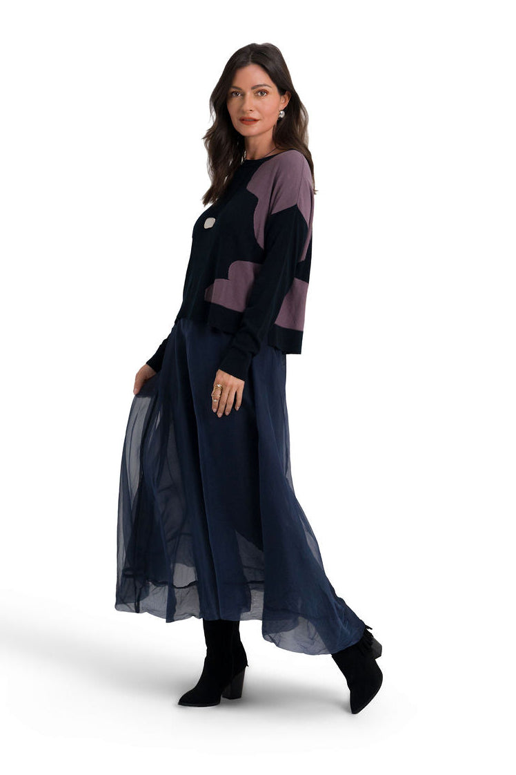 A woman modeling a long-sleeved organza skirt with a color-blocked pima cotton sweater in navy blue, black, and muted purple, paired with black ankle boots, against a plain white studio background.