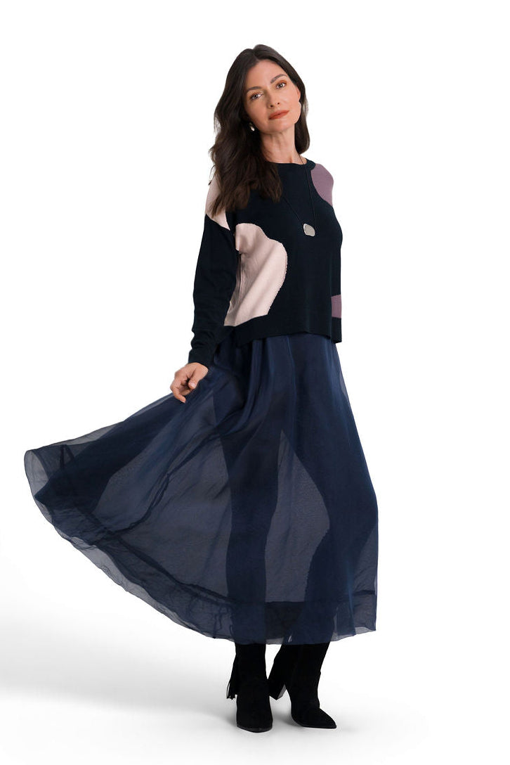 A woman modeling a long midnight navy blue organza skirt and contrasting light panels on the sleeves of a pima cotton sweater, standing against a solid white studio background.
