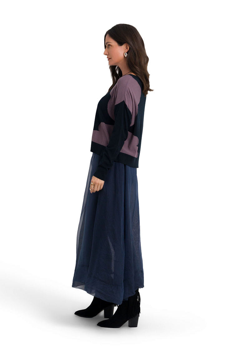 A woman modeling a long-sleeve color-blocked pima cotton sweater and matching maxi skirt set. The set features dark navy blue and muted purple color blocking, styled with black ankle boots against a plain white studio background.