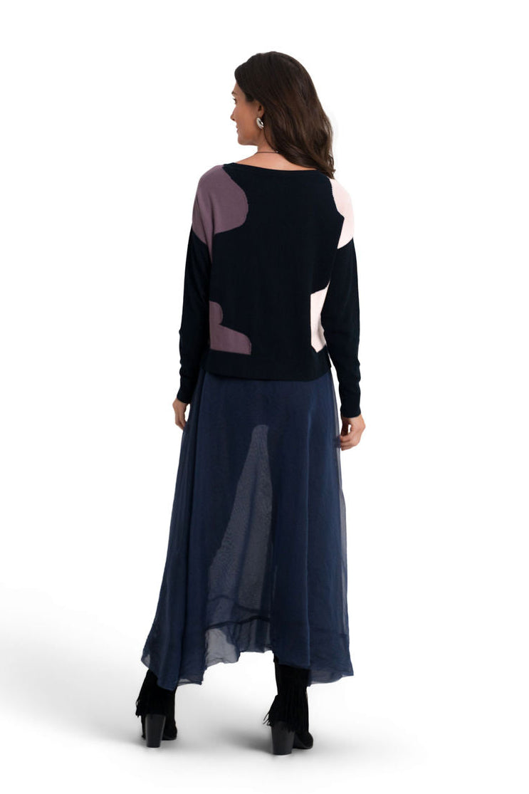 A woman is shown from the back wearing a long-sleeve pima cotton sweater with abstract color-blocked panels in black, dusty purple, and off-white, paired with a long midnight navy blue skirt and black boots, against a plain white background.